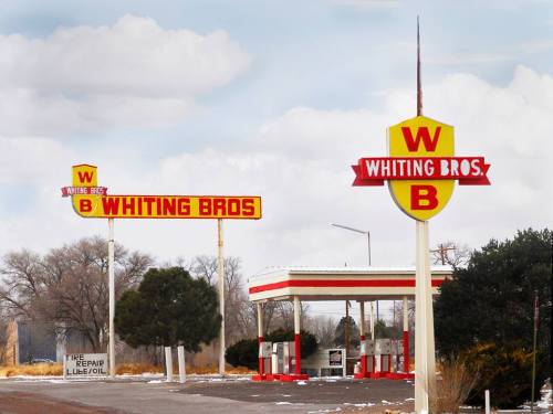A vintage gas station with "Whiting Bros" signage, surrounded by sparse trees and a cloudy sky.