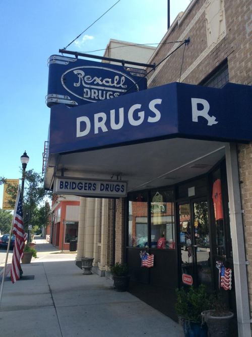 A vintage drugstore with a blue sign reading "Rexall Drugs" and "Bridgers Drugs," featuring American flags outside.