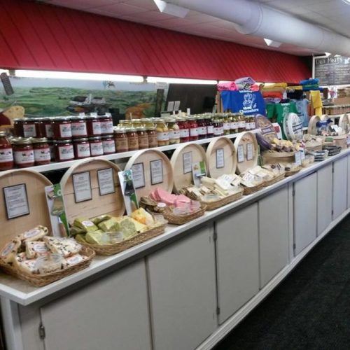 A display of various food products, including jars, cheeses, and snacks, in a market setting with colorful shirts in the background.