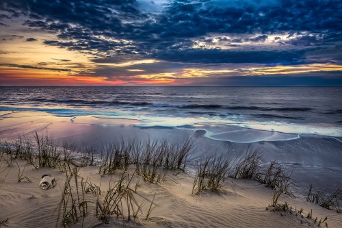 A serene beach at sunset, with gentle waves, sandy dunes, and grass under a colorful sky filled with clouds.