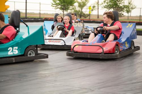 A group of people racing go-karts on a track, with colorful karts in motion and trees in the background.