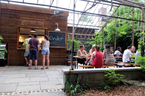 A cozy outdoor café with people ordering at a counter and others seated at tables surrounded by greenery.