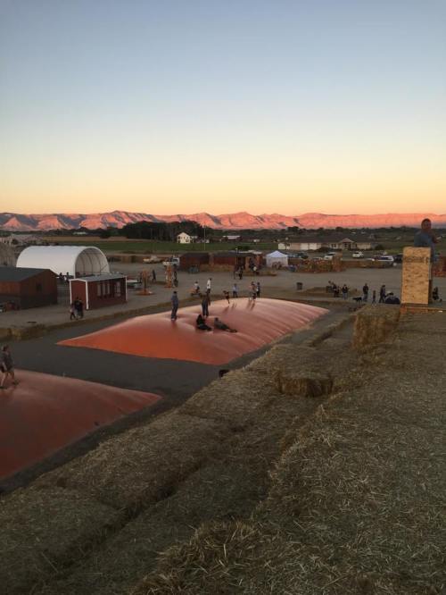 Children and adults play on large inflatable surfaces at sunset, with mountains in the background and hay bales nearby.