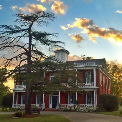 A historic red brick building with white trim, surrounded by trees and blooming flowers, under a colorful sunset sky.