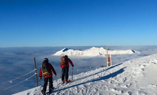 Two climbers in red jackets stand on a snowy peak, overlooking a sea of clouds and distant mountains under a clear blue sky.
