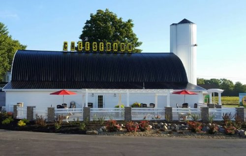 A white barn-style building with a black roof, featuring a sign that reads "Cheese Barn" and red umbrellas outside.