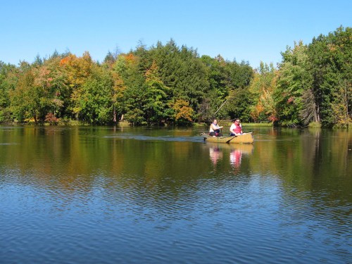 Two people in a canoe fishing on a calm lake surrounded by colorful autumn trees under a clear blue sky.