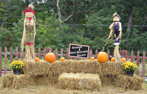 Two scarecrows stand beside pumpkins and hay bales, with a sign for Riverside & Great Northern Railway in the background.