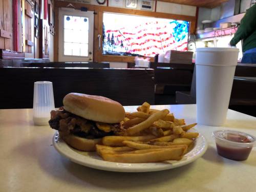 A burger with bacon and cheese, fries, and a drink on a table in a cozy diner with an American flag backdrop.