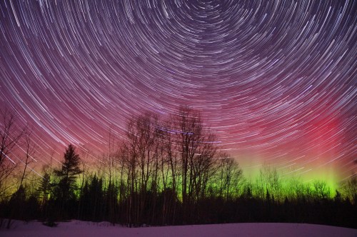 A starry night sky with swirling star trails and vibrant green and red auroras above a snowy landscape and trees.