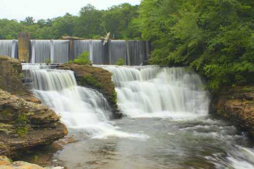 A serene waterfall cascades over rocky ledges, surrounded by lush green trees and a tranquil river.