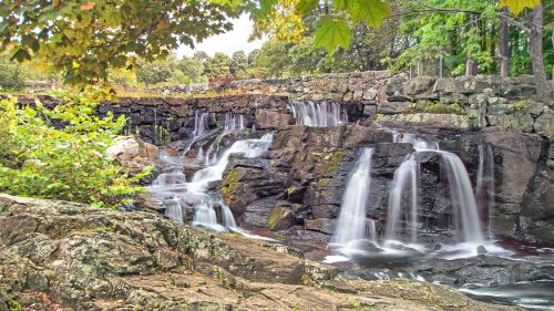 A serene waterfall cascades over rocky ledges, surrounded by lush greenery and autumn foliage.