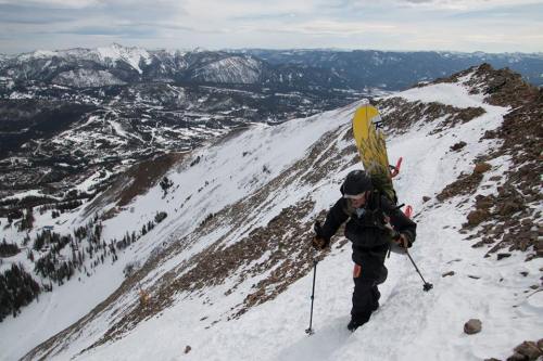 A person climbs a snowy mountain slope, carrying a snowboard and gear, with a scenic view of mountains in the background.