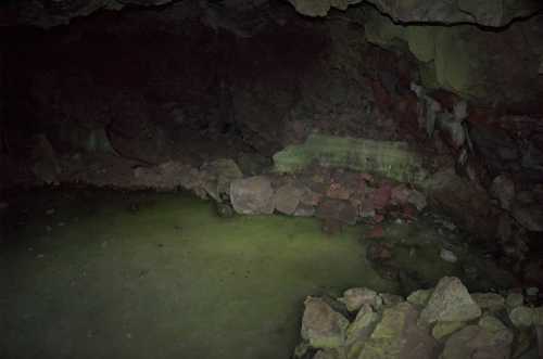 A dark cave interior with a small pool of water and scattered rocks on the ground.