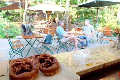 Two people chat at an outdoor café with tables and umbrellas, while pretzels sit on a counter in the foreground.