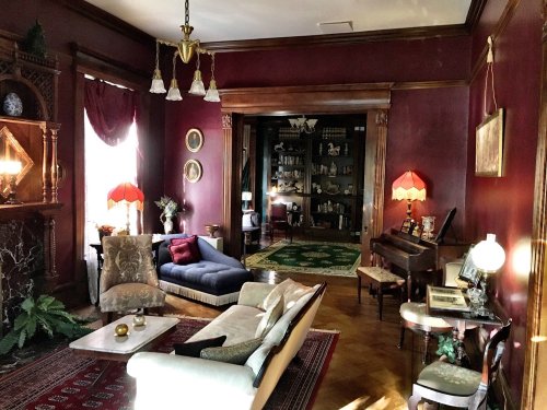 A cozy, vintage living room with rich red walls, elegant furniture, and warm lighting, featuring a piano and decorative accents.