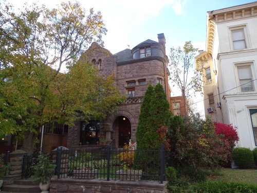 A historic stone house with a turret, surrounded by trees and colorful shrubs, next to a lighter-colored building.