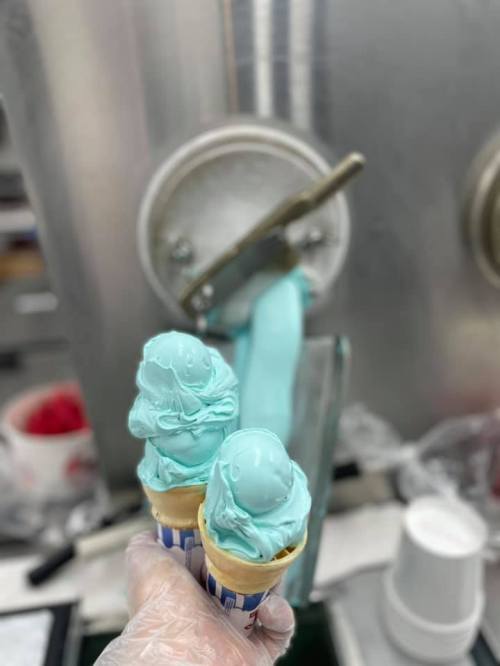Two ice cream cones with blue mint ice cream, held in a gloved hand, with a soft serve machine in the background.