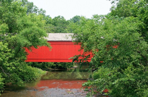 A red covered bridge spans a calm stream, surrounded by lush green trees and foliage.