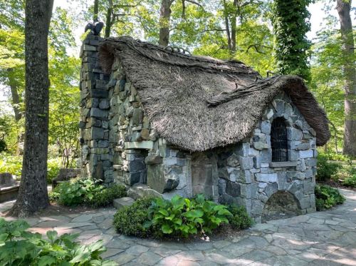 A quaint stone cottage with a thatched roof, surrounded by lush greenery and trees.
