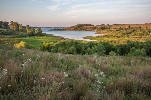 A serene landscape featuring a lake surrounded by green hills and lush vegetation under a clear sky.