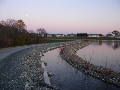 A winding path beside a calm waterway at dusk, with a full moon and houses in the background.