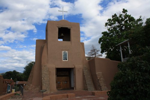 Adobe-style church with a cross on top, bell tower, and surrounding greenery under a partly cloudy sky.
