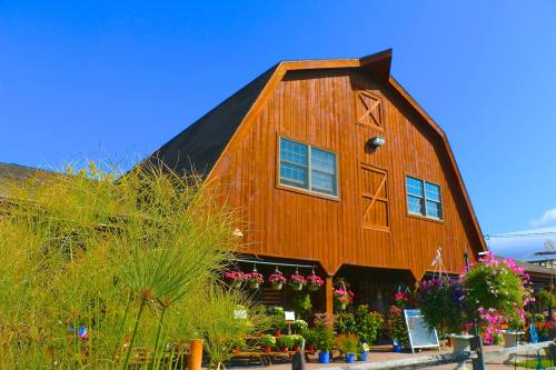 A large wooden barn-style building with windows, surrounded by colorful flowers and greenery under a clear blue sky.