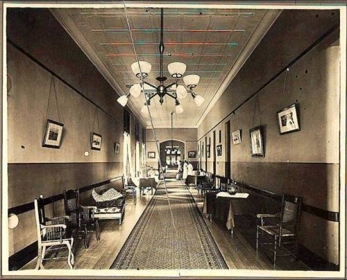 A vintage hallway with wooden floors, chandeliers, and framed pictures on the walls, leading to a doorway.