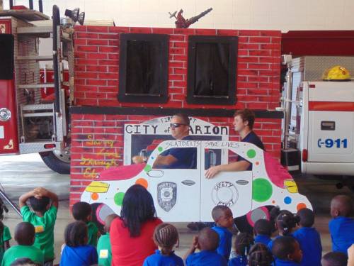 Children watch as two adults demonstrate safety in front of a colorful fire truck and a painted car backdrop.