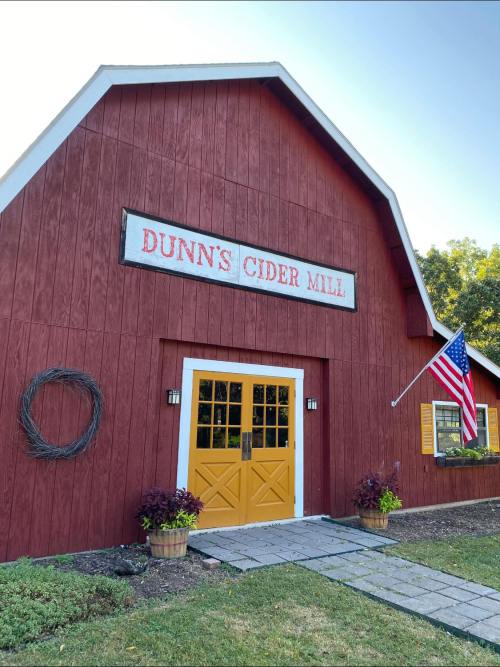 Red barn with a sign reading "Dunn's Cider Mill," yellow doors, American flag, and decorative plants at the entrance.