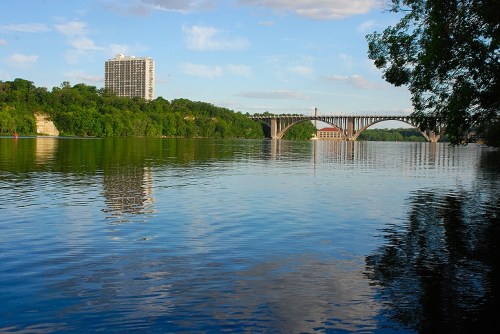 A serene river scene with a bridge, lush greenery, and a building reflected in the calm water under a blue sky.