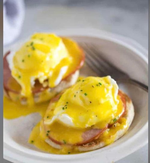 Two eggs Benedict on English muffins, topped with hollandaise sauce and garnished with chives on a white plate.