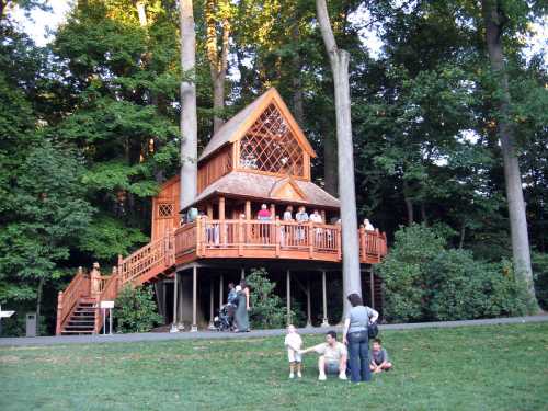 A large wooden treehouse surrounded by trees, with people on the deck and children playing on the grass below.