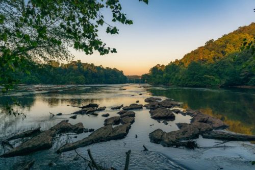 A serene river scene at sunset, with rocky outcrops and lush green hills reflecting in the calm water.