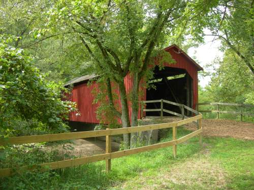 A red covered bridge nestled among green trees, with a wooden fence and a path leading to it.