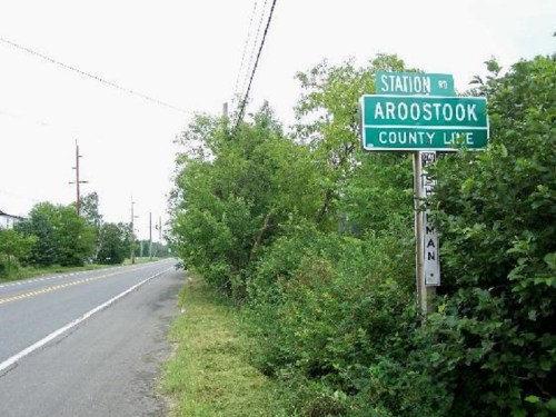 A roadside sign indicating the Aroostook County line, surrounded by greenery and a quiet road.