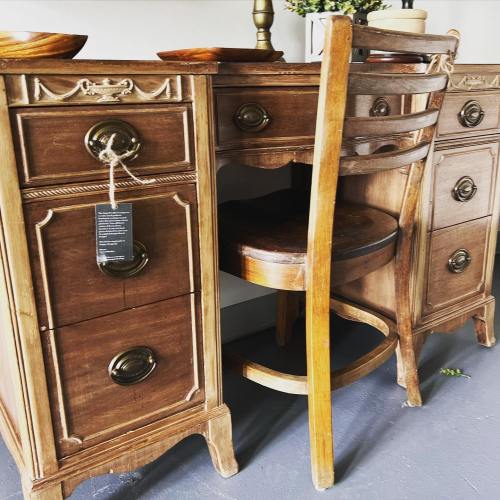 A wooden vintage-style desk with drawers and a chair, set against a neutral background.