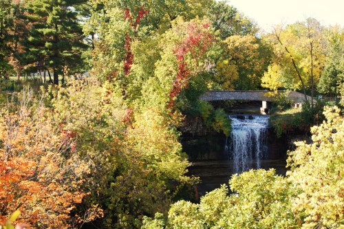 A scenic view of a waterfall surrounded by vibrant autumn foliage and a stone bridge above.