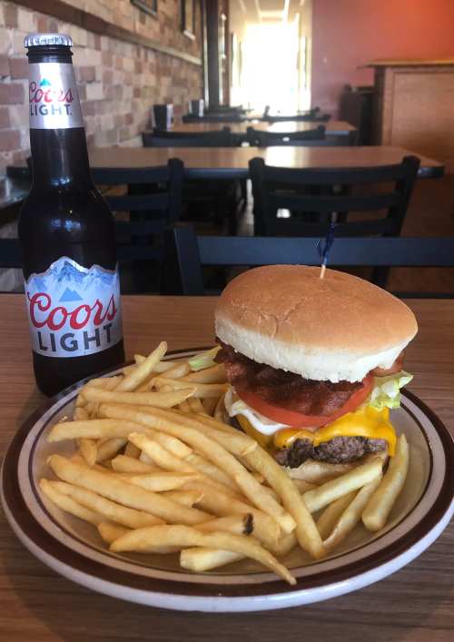 A cheeseburger with lettuce, tomato, and bacon, served with fries and a bottle of Coors Light in a casual dining setting.