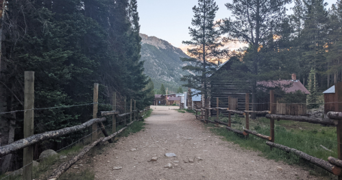 A dirt path lined with wooden fences leads through trees to rustic buildings and mountains in the background.