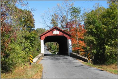 A red covered bridge spans a road, surrounded by trees with autumn foliage under a clear blue sky.