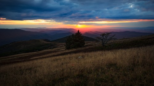 A sunset over rolling mountains, with a grassy foreground and scattered trees under a dramatic sky.