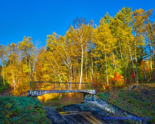 A concrete bridge over a dry creek, surrounded by vibrant autumn trees and a clear blue sky.
