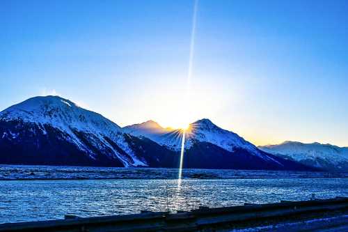 Sunrise over snow-capped mountains, reflecting on a calm body of water, with a clear blue sky above.