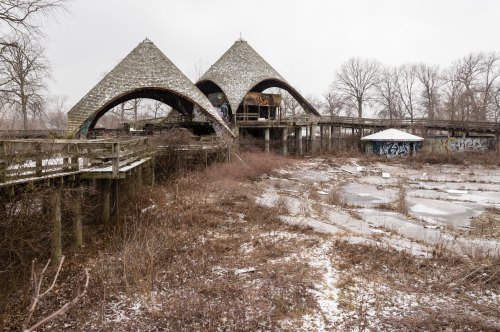Abandoned building with unique triangular roofs, surrounded by overgrown vegetation and remnants of a frozen landscape.