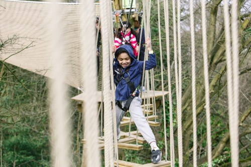 A child joyfully navigates a rope bridge, wearing a harness and smiling, surrounded by trees.