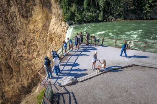 A group of people stands on a viewing platform near a river with a waterfall, surrounded by lush greenery and rocky cliffs.