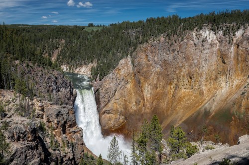 A stunning waterfall cascades down a rocky cliff surrounded by lush green trees and a clear blue sky.