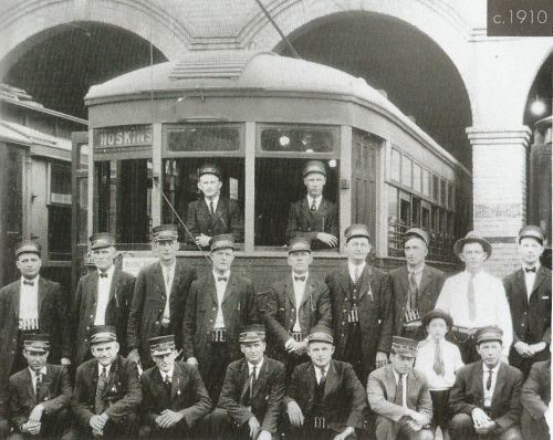 Black and white photo of a group of men in uniforms posing in front of a vintage streetcar, circa 1910.